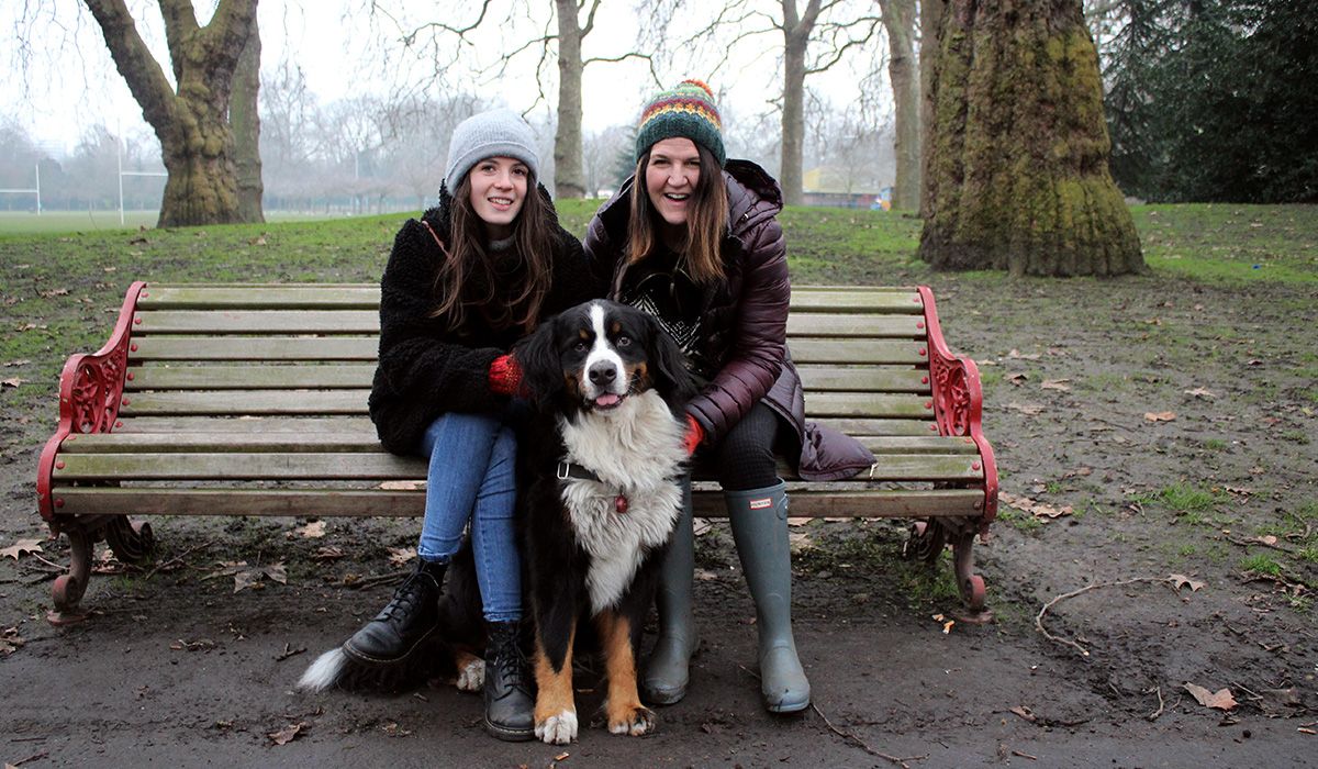 Two women sit on a park bench, smiling. Sitting between them is a large dog.