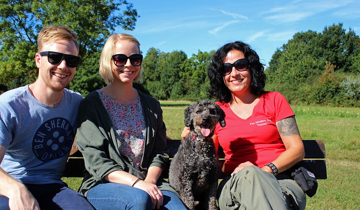 Three people and a black, curly-haired dog are sitting on a bench in a park